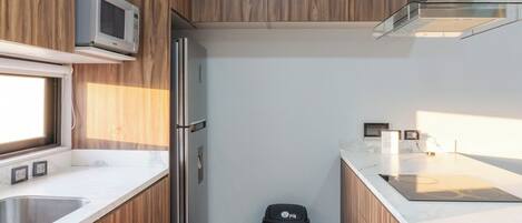 Una cocina compacta con armarios de madera, un refrigerador de acero inoxidable, un microondas y un cubo de basura.