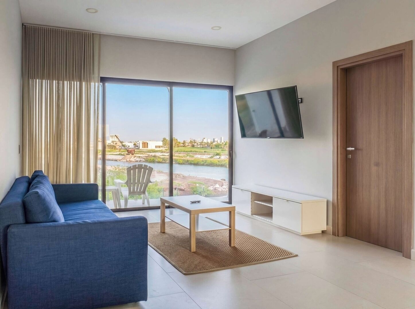 Una sala moderna con un sofá, una mesa, un televisor y un ventanal corredera que da a vistas de un río y edificios.