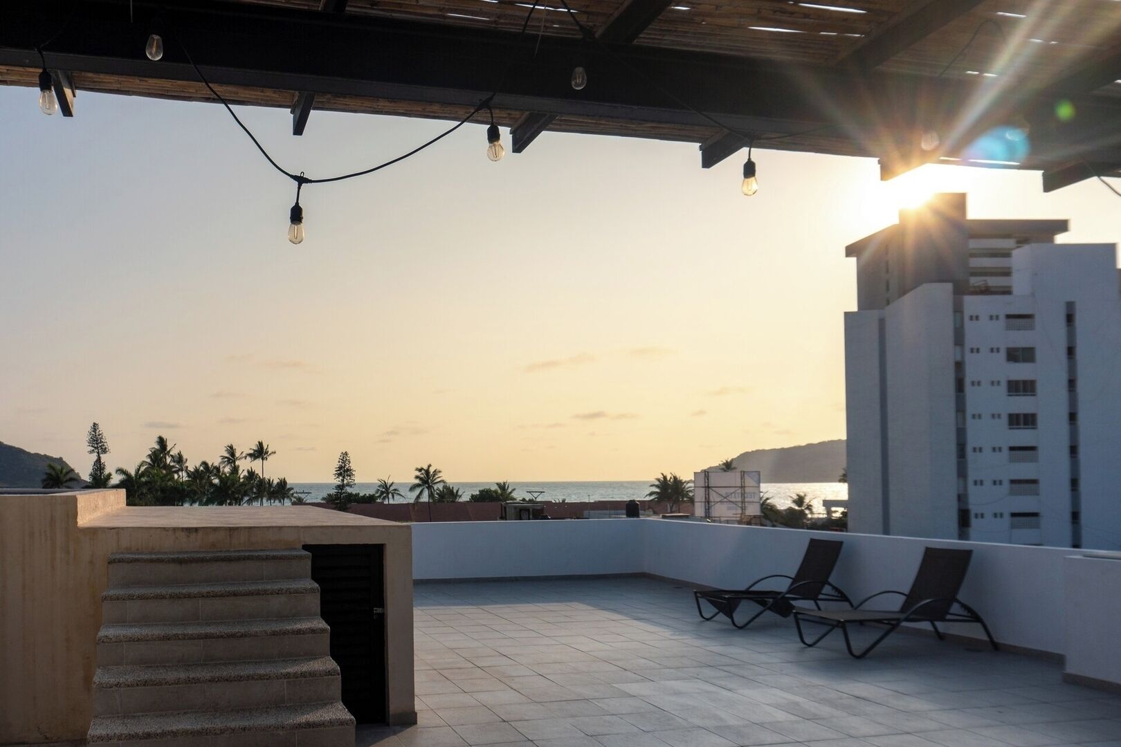 Eine Dachterrasse mit Blick auf das Meer, einem Holztisch, zwei Lounge-Stühlen und einem modernen Gebäude im Hintergrund.