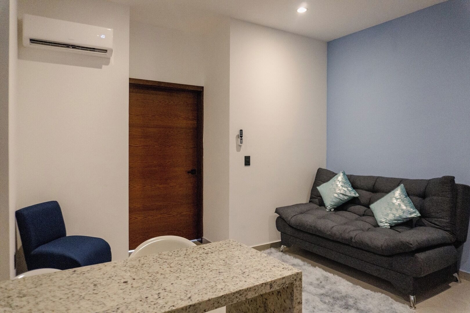 Ein Zimmer mit einem dunkelgrauen Sofa, einem kleinen Tisch, einem blauen Stuhl und einer Holztür.