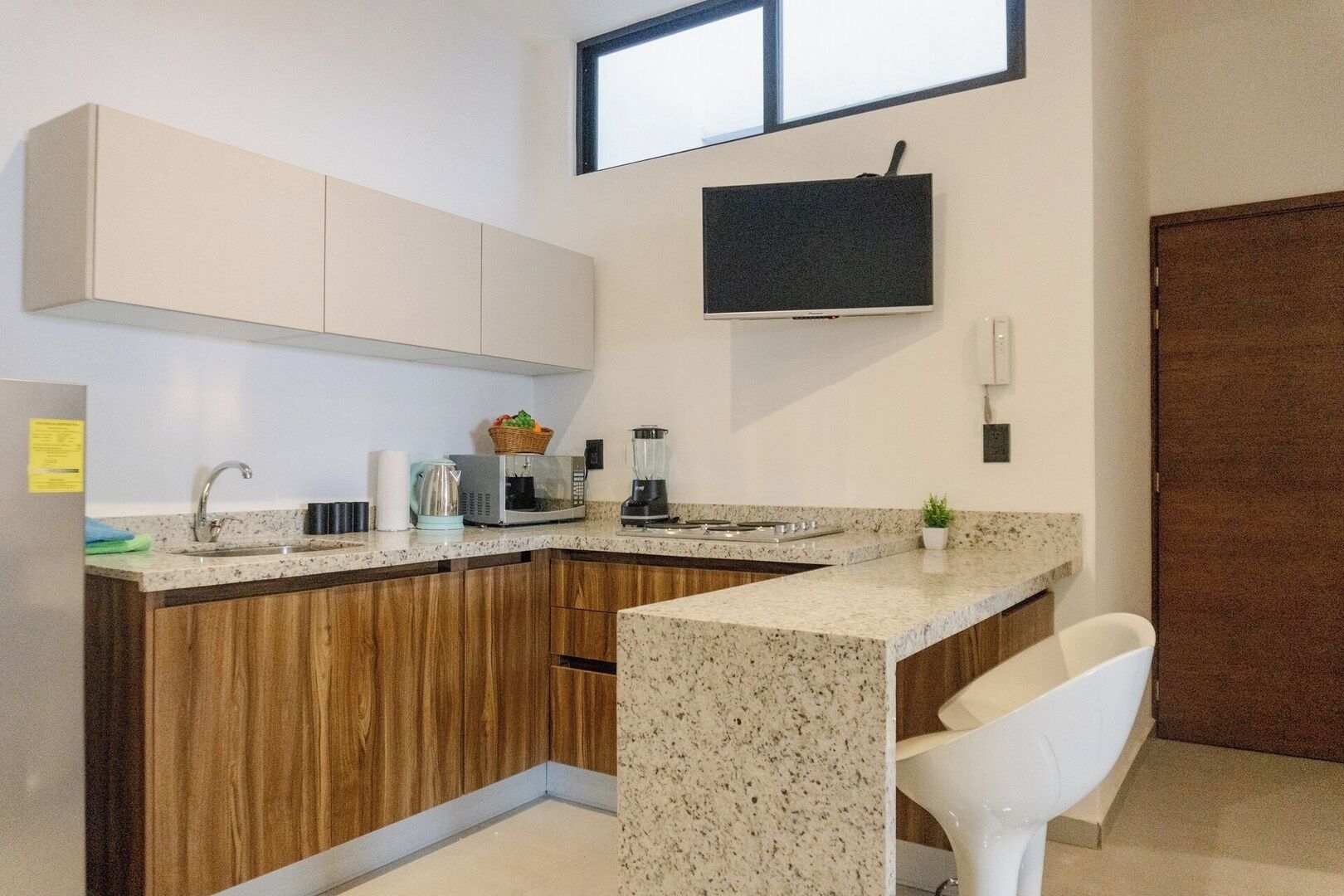 Une cuisine équipée d’un four à micro-ondes, d’une bouilloire et d’un mixeur sur le plan de travail, de placards en bois, d’une chaise blanche et d’un téléviseur fixé au mur.