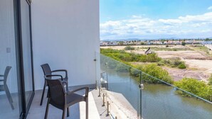 Un balcon avec deux chaises, une rambarde en verre et une vue sur une rivière et des bâtiments éloignés.