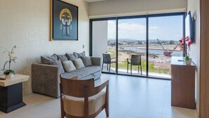 Un salon moderne comprenant un canapé, des fauteuils, une table basse et une grande fenêtre donnant sur un plan d’eau et sur des bâtiments situés au loin.