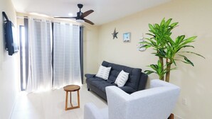 Ein Wohnzimmer mit einer Couch, einem Sessel, einem kleinen Holzhocker, einem Deckenventilator, einer Topfpflanze und einer Sternen-Dekoration an der Wand.
