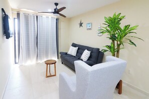 Ein Wohnzimmer mit einer Couch, einem Sessel, einem kleinen Holzhocker, einem Deckenventilator, einer Topfpflanze und einer Sternen-Dekoration an der Wand.