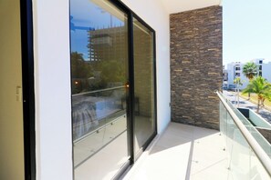 Een balkon met een glazen deur, een stenen muur en uitzicht op een straat met gebouwen en palmbomen.