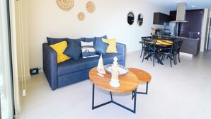 Ett modernt vardagsrum med en blå soffa, gula kuddar, ett träbord och en matplats med ett kök synligt i bakgrunden.