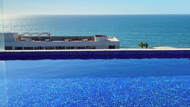 En pool med blå kakelplattor, en byggnad med vit fasad och utsikt över havet.