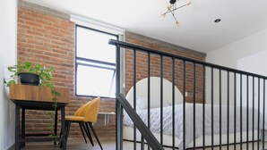 Un dormitorio con pared de ladrillo, una cama con cabecera blanca, un escritorio con planta, una silla amarilla y una barandilla negra.