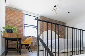 Ein Schlafzimmer mit Backsteinwand, einem Bett mit weißem Kopfteil, einem Schreibtisch mit Pflanze, einem gelben Stuhl und einem schwarzen Geländer.