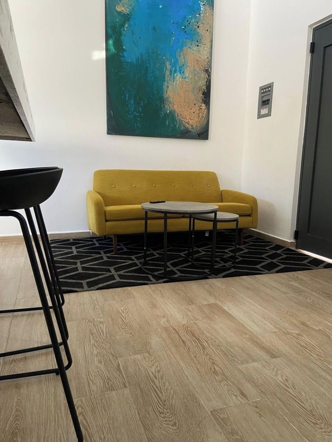 Une pièce avec un canapé jaune, une petite table ronde, une chaise en métal noir, un tapis à motifs géométriques et un grand tableau abstrait accroché au mur.