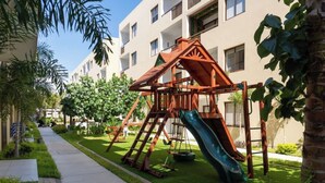 Un parque infantil con un tobogán y columpios, rodeado de un césped cuidado y una acera.