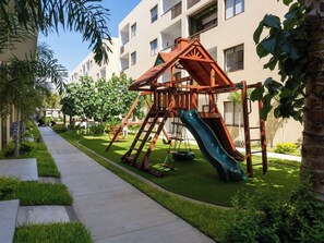 Ein Spielplatz mit Rutsche und Schaukeln, umgeben von einem gepflegten Rasen und einem Bürgersteig.