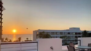 Een balkon met een tafel en stoelen, uitkijkend op een zonsondergang boven de oceaan, met een gebouw op de achtergrond.
