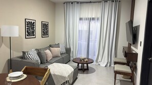 Ein modernes Wohnzimmer mit einem grauen Sofa, einem kleinen runden Tisch, einem an der Wand montierten Fernseher und einem Fenster mit transparenten Vorhängen.