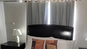 A hotel room with a bed, a nightstand with a lamp, a wall-mounted air conditioner, and a window with curtains. - Stylish apartment close to attractions, dining, shopping, and entertainment (Montego Bay)