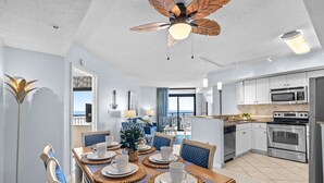 Una cocina moderna con electrodomésticos de acero inoxidable, un comedor con mesa de madera y sillas azules, y un ventilador de techo con diseño de hojas.