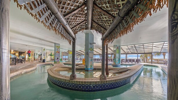 Una piscina interior con borde de azulejos, una fuente central y una estructura de techo de paja.