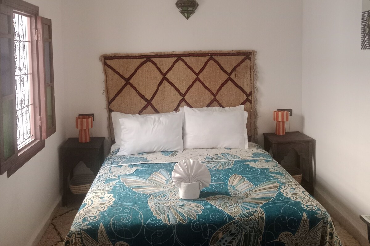 A bed with a patterned bedspread, two pillows, a decorative wall hanging, and a ceiling light fixture.
