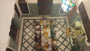 A dining area with a long table surrounded by chairs, a patterned floor, and a stained glass window above.