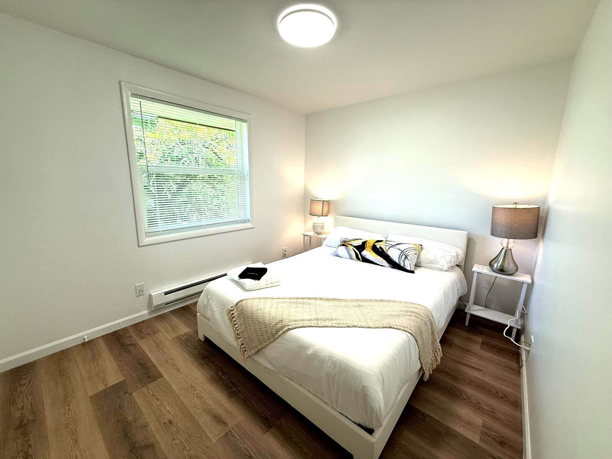 Une chambre à coucher avec un lit, deux lampes de chevet, une fenêtre avec des stores, un radiateur et un luminaire au plafond.
