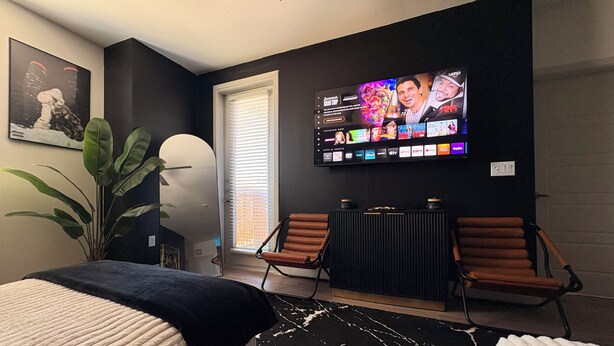 Een moderne hotelkamer met een grote flatscreen-televisie aan de muur, een bed met een zwart-wit vloerkleed, twee bruine stoelen en een potplant.