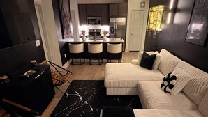 Een moderne woonkamer met een bank, een salontafel en een televisie. Achtergrond toont een keuken met een eettafel en stoelen.