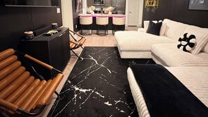 Een moderne woonkamer met een bank, een zwart-wit kleed, een houten stoel en een keuken in de achtergrond.