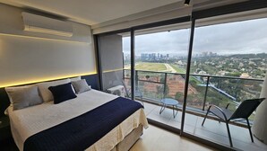 A modern bedroom with a large bed, a view of a cityscape, and a balcony with chairs. - Studio Espaçoso - Pinheiros (São Paulo)
