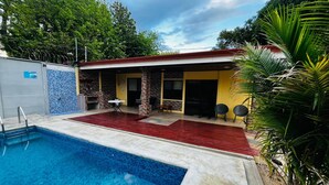Une piscine bordée d’une terrasse rouge, un patio couvert avec des chaises et un mur en pierre.