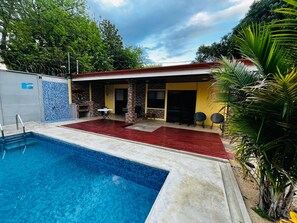 A swimming pool with a red deck, a covered patio with chairs, and a stone wall. - Casa Del Sol: Privacy, comfort and unforgettable moments. (Provincia de Guanacaste)
