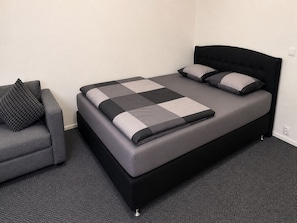 A single bed with a black frame and grey bedding, a grey armchair with a black cushion, and a plain white wall. - Nordstay Turku (Turku)