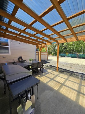 Ein überdachter Patio-Bereich mit Tisch, Stühlen, einem Grill und einer Pergola aus Holz.