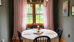Une salle à manger avec une table ronde, des chaises, un lustre et une vue sur une maison, visible à travers de grandes fenêtres.