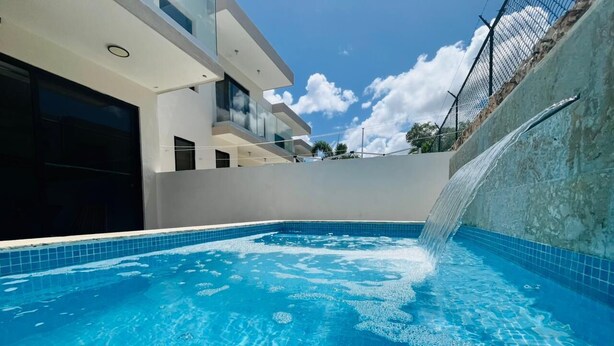 A swimming pool with a waterfall feature, surrounded by a modern building with balconies. - 3-bedroom house in tranquil Punta Cana with cool AC breeze (Punta Cana)