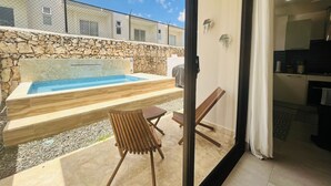 A modern outdoor pool area with a stone wall, wooden deck, and a glass door leading to a kitchen. - 3-bedroom house in tranquil Punta Cana with cool AC breeze (Punta Cana)