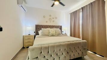 A bedroom with a tufted bed, two bedside tables with lamps, a ceiling fan, and a window with curtains.