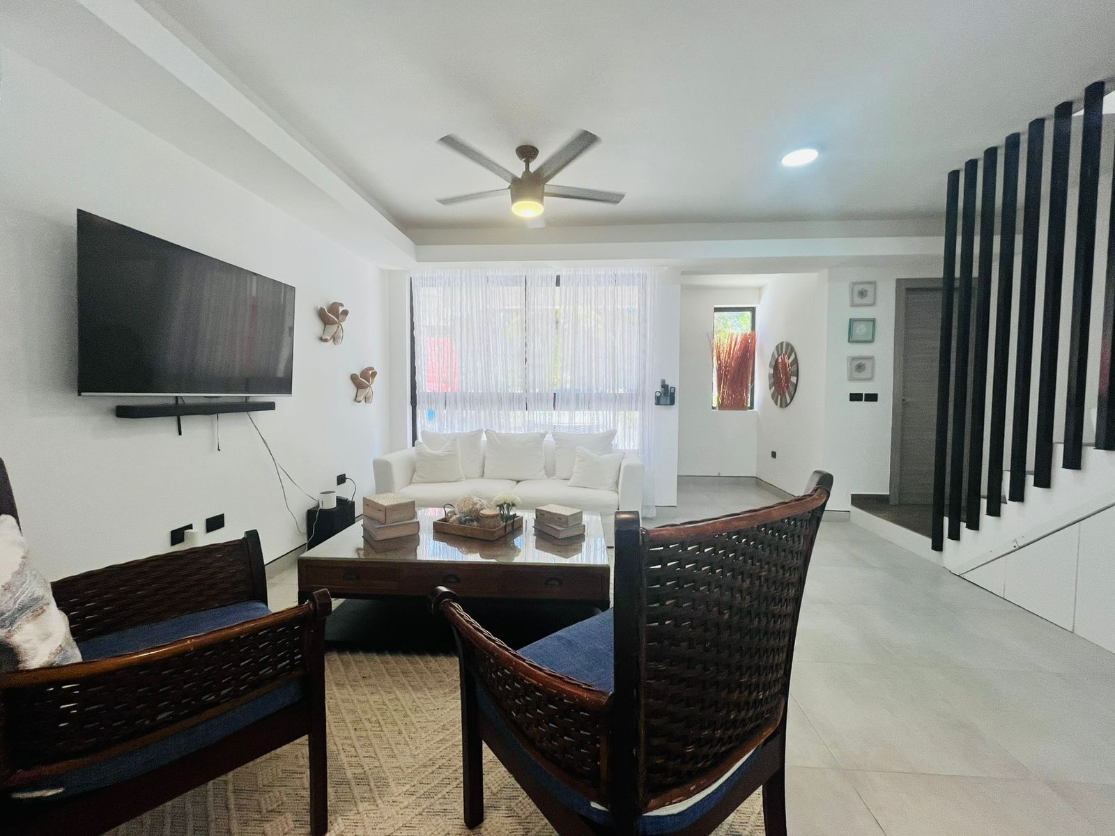 A modern living room with a flat-screen TV, a ceiling fan, a sofa, a coffee table, and a staircase with black railings.