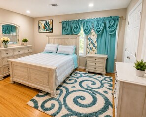 Une chambre à coucher comprenant un lit, une commode, une table de chevet et une grande fenêtre avec des rideaux.