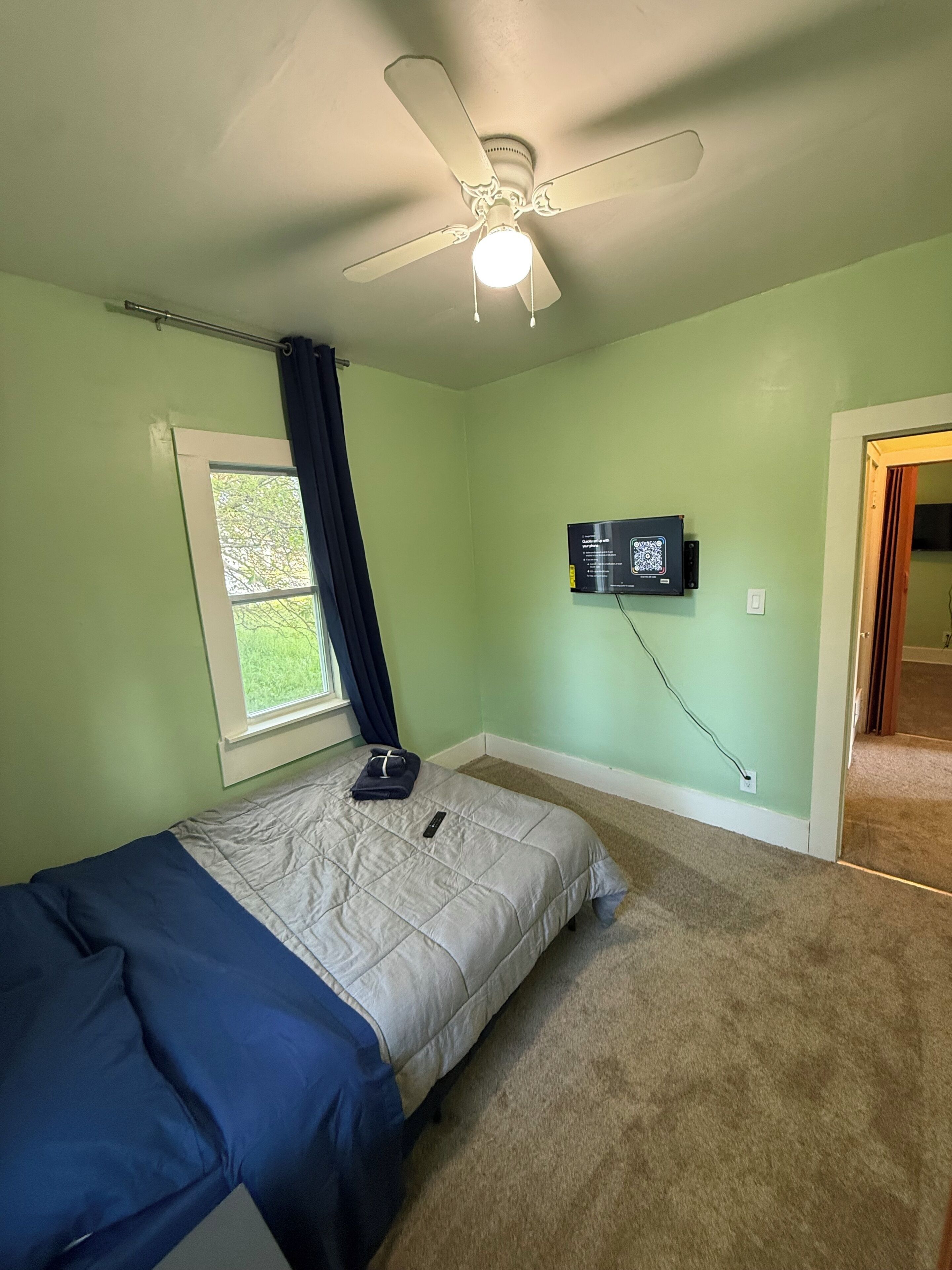 Une chambre à coucher comprenant un lit, un ventilateur de plafond, une fenêtre avec des rideaux, un téléviseur fixé au mur et un sol recouvert de moquette.