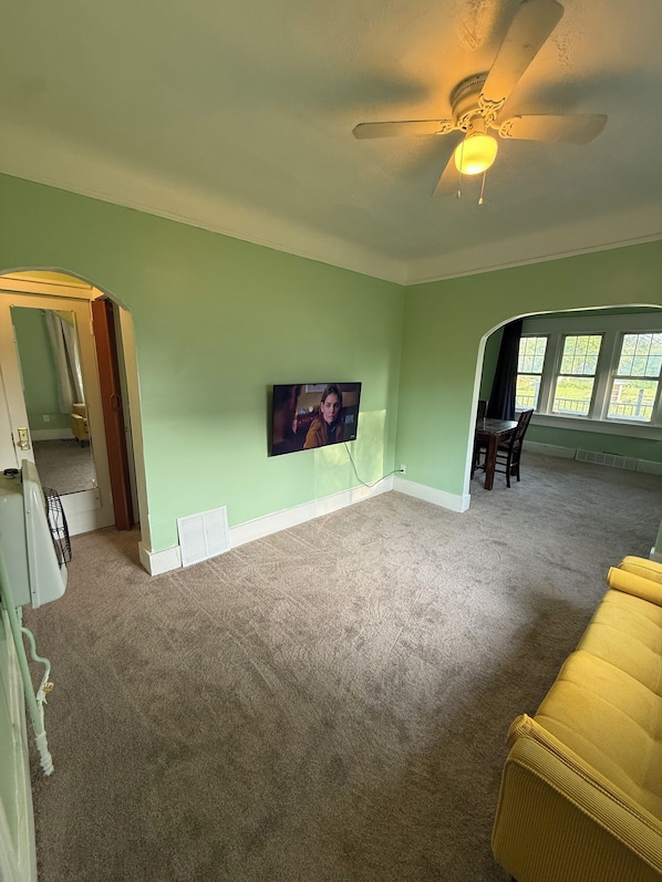 Un salon spacieux doté d’un téléviseur à écran plat fixé au mur, d’un ventilateur de plafond et d’un canapé jaune.