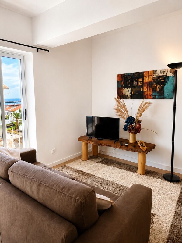 Ein modernes Wohnzimmer mit einer Couch, einem Fernseher auf einem Holzboden, einem Bild an der Wand und Blick durch eine Glastür.