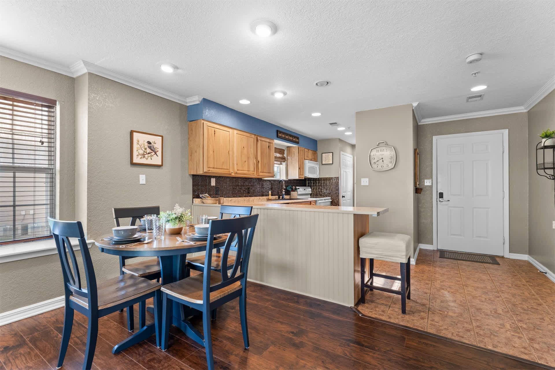 A kitchen with wooden cabinets, a dining table with chairs, a clock on the wall, and a door with a mat.