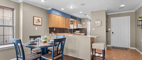 A kitchen with wooden cabinets, a dining table with chairs, a clock on the wall, and a door with a mat.