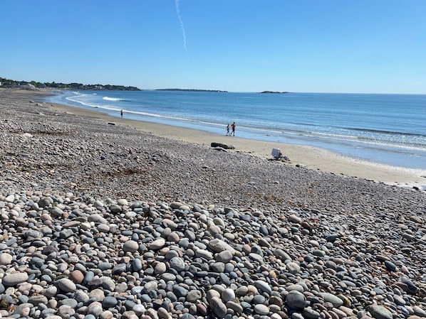 A pebbled beach with a clear sky, distant shoreline, and a few people walking.