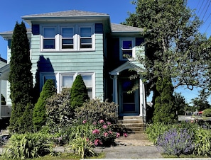 A two-story house with a front porch, a small garden with flowers, and a tall tree.