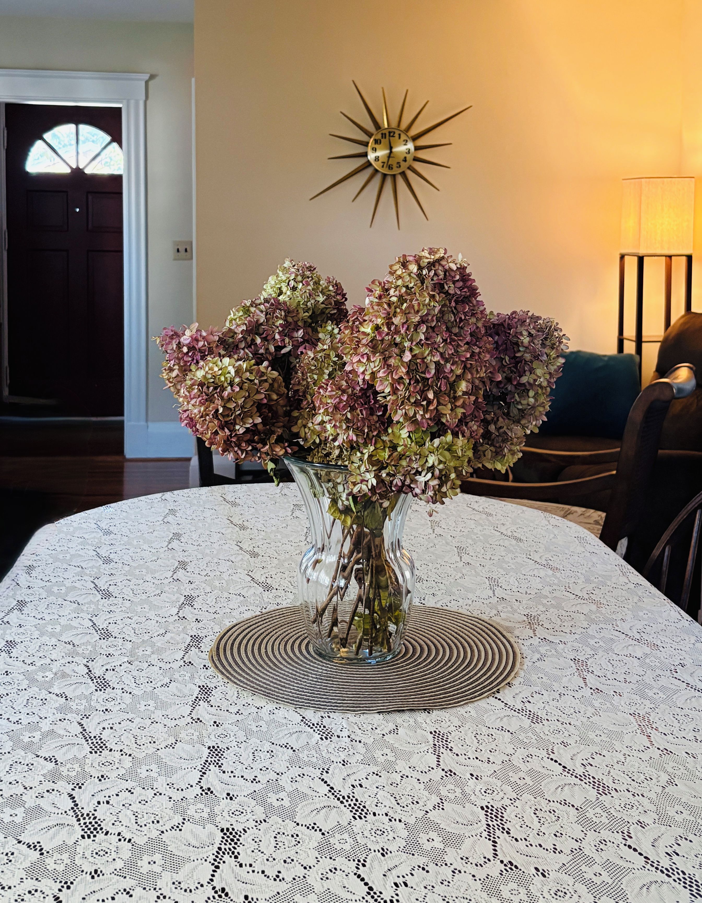 A table with a lace tablecloth, a vase with flowers, and a sunburst wall decoration.