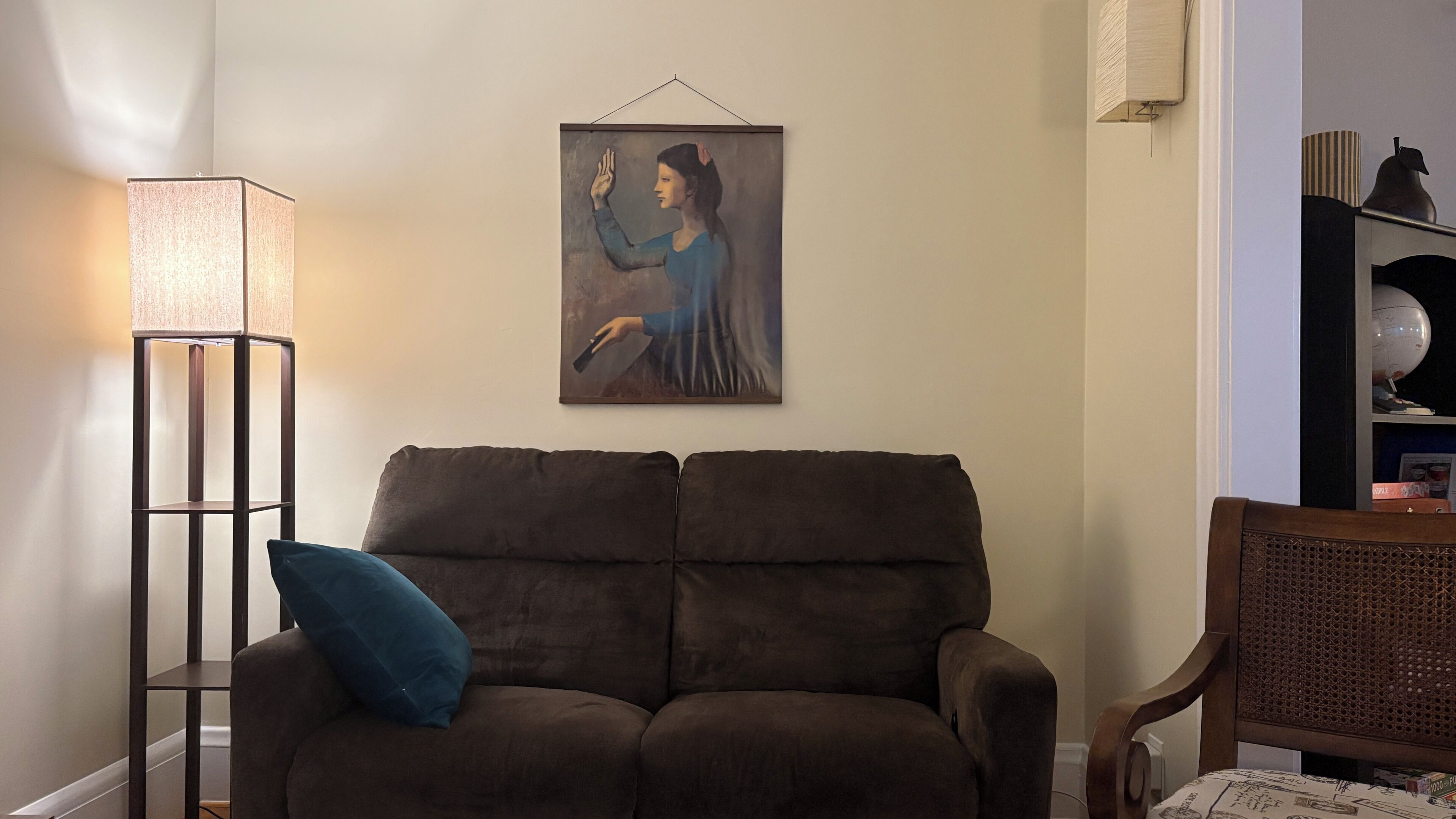 A living room with a brown sofa, a blue pillow, a floor lamp, and a painting on the wall.