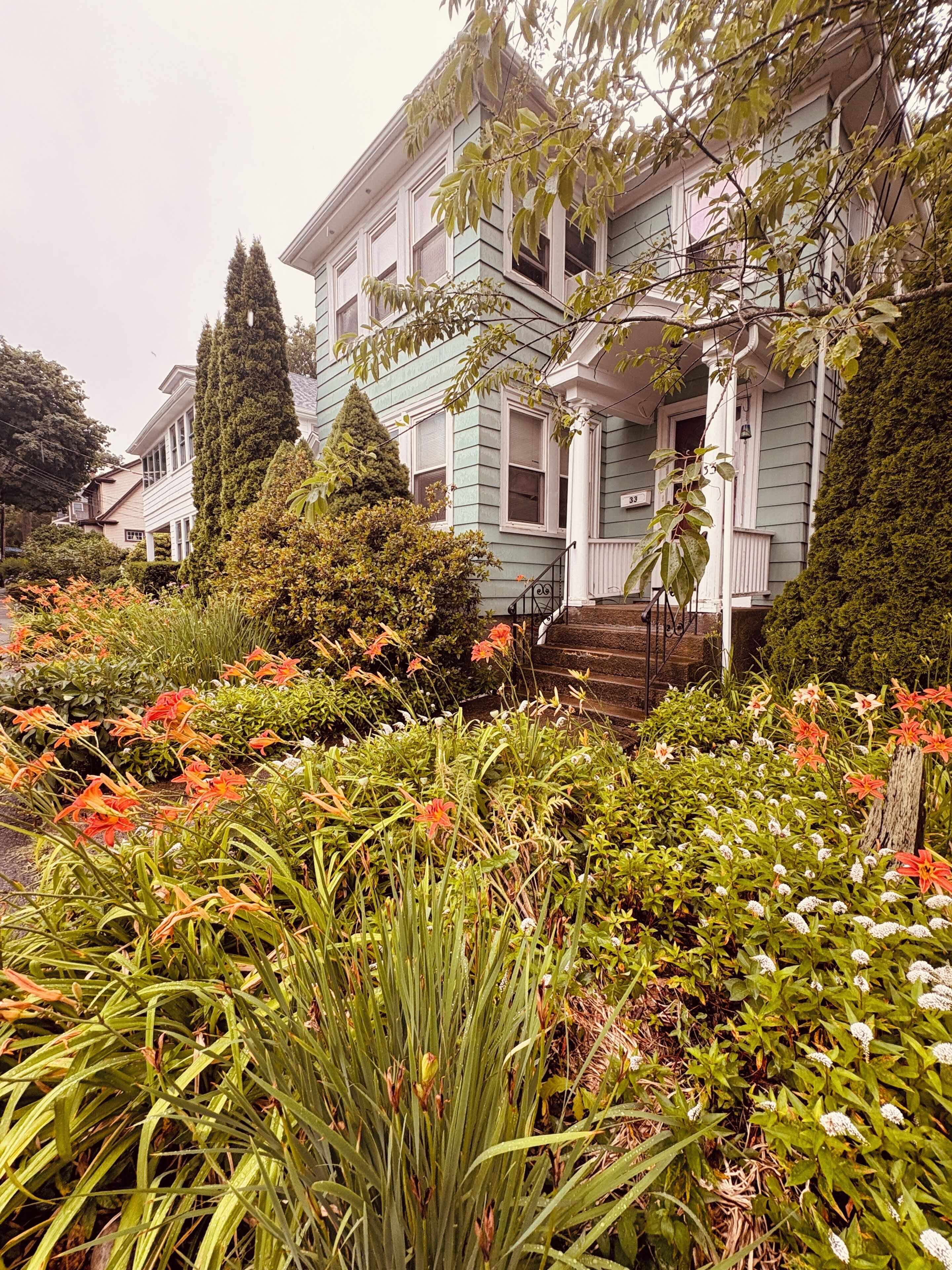 A two-story house with a front porch, surrounded by a variety of plants and flowers.
