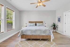 Ein Schlafzimmer mit einem Bett, einem Deckenventilator, einem Nachttisch, einer Topfpflanze und einem Fenster mit Jalousien.